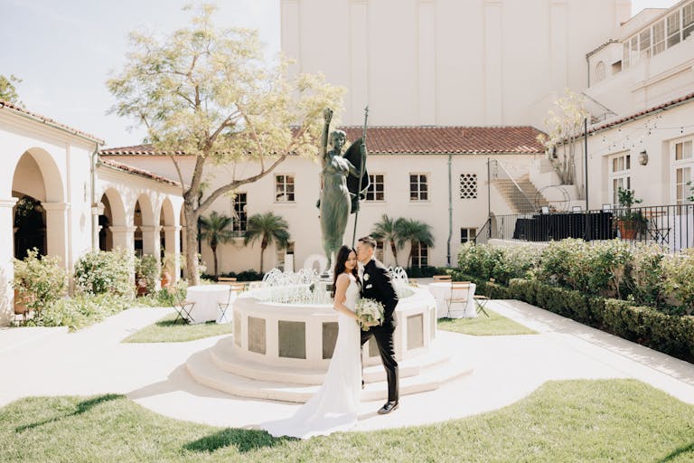 Groom kisses bride's templet in front of fountain in The Courtyard Garden at The Ebell of Los Angeles | PartySlate