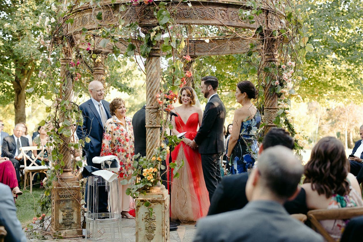 Bride wears red dress at ceremony under a garden gazebo | PartySlate