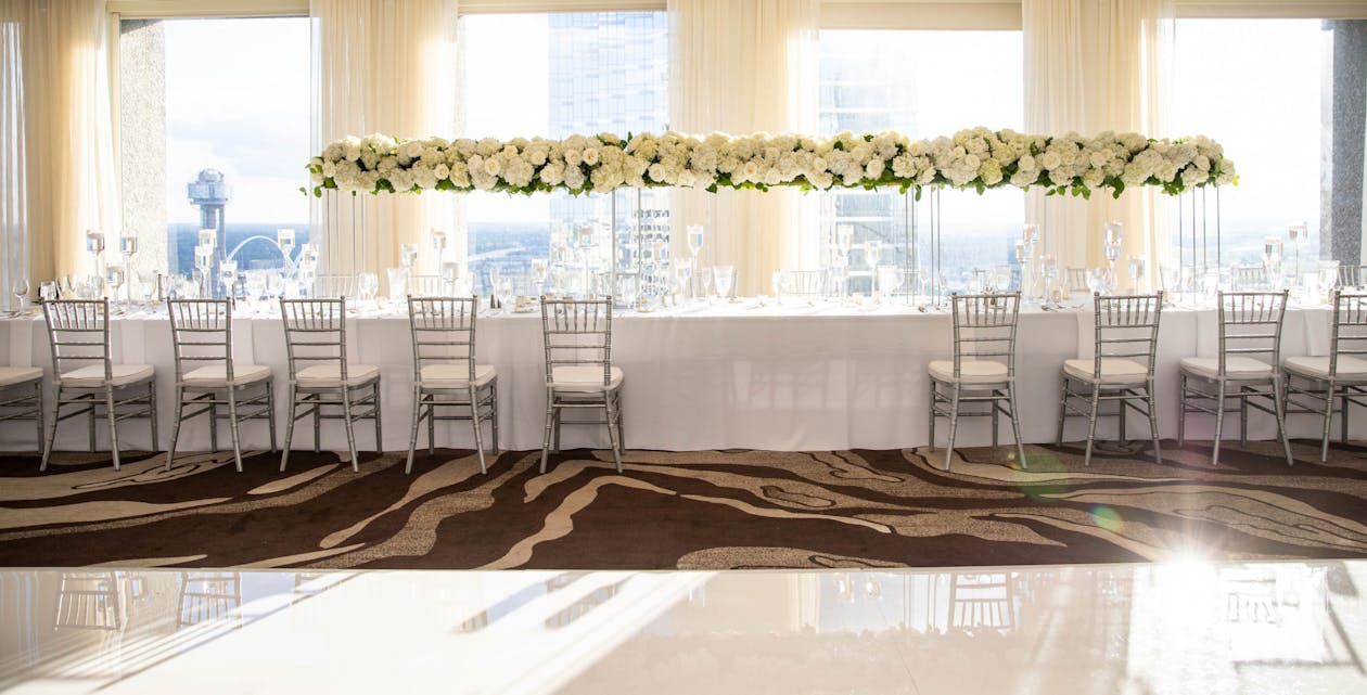 head table at reception with raised centerpiece of white florals and greenery with a white table cloth on the table | PartySlate