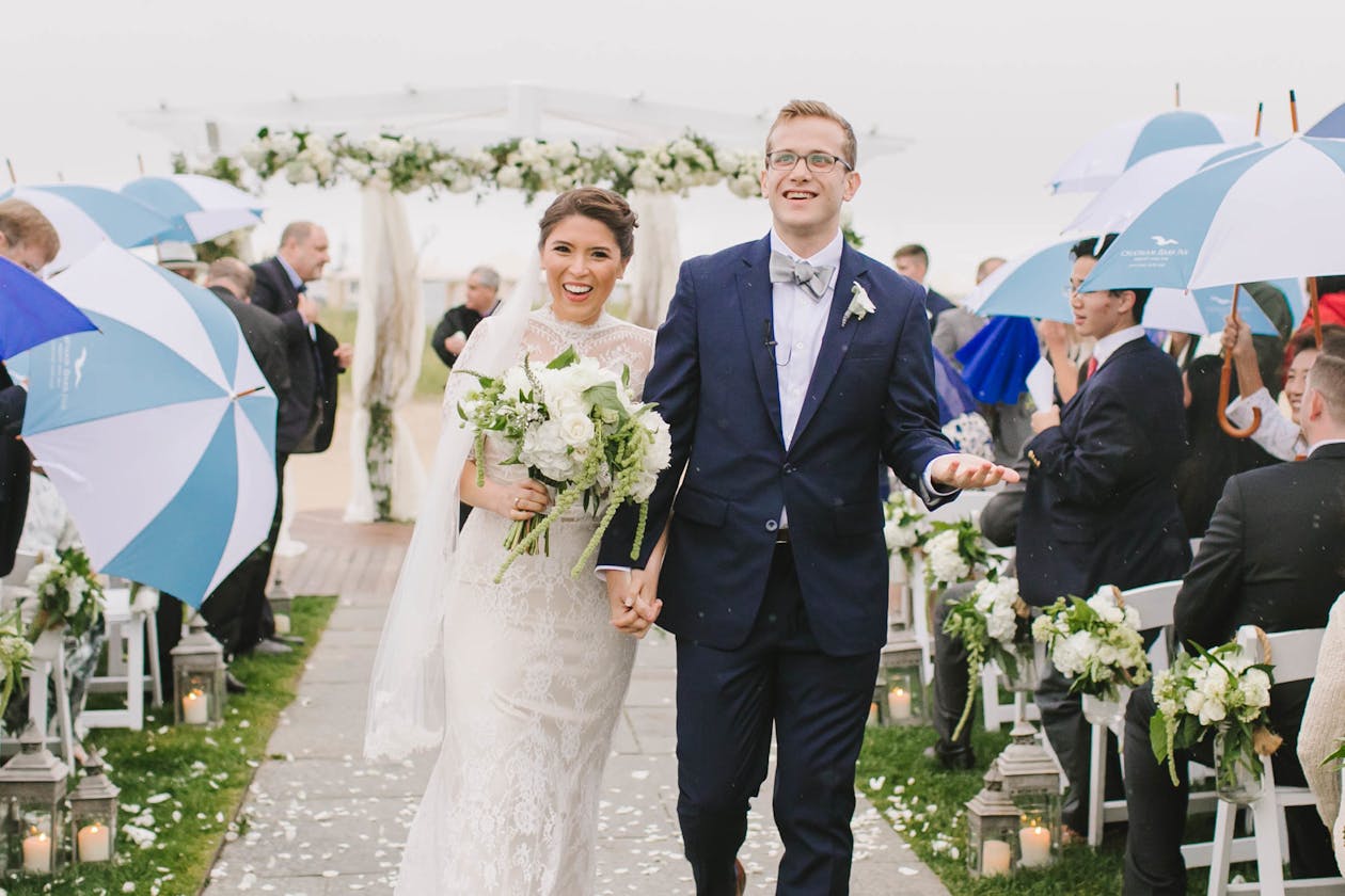 Couple walks down wedding aisle while guests hold blue and white umbrellas | PartySlate
