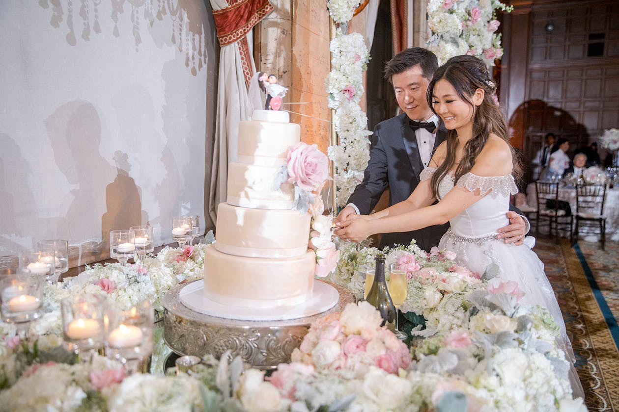 Couple cut white wedding cake surrounded by pink and white flowers | PartySlate