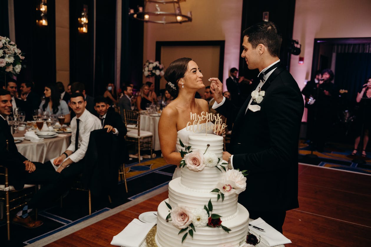 White three tiered classic wedding cake with florals on it and couple tasting the cake | PartySlate
