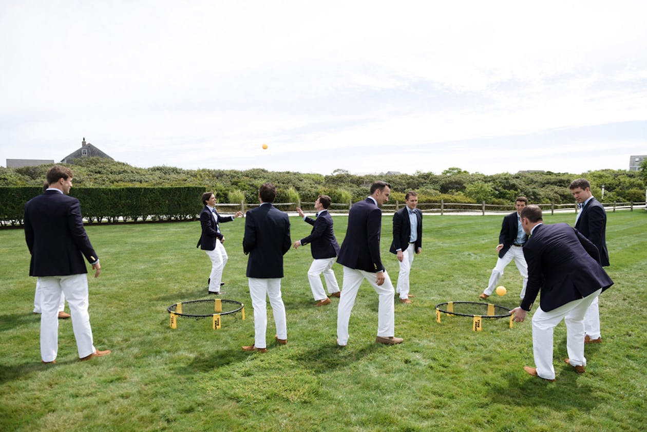 Preppy groomsmen in blue jackets and white pants play spikeball on green lawn | PartySlate