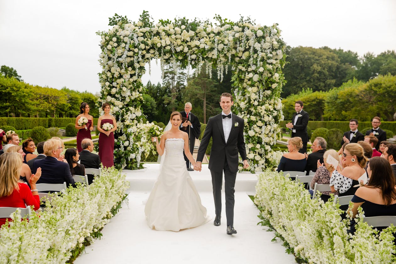 Floral squared arch outside covered in greenery ad white florals with couple walking down the aisle | PartySlate