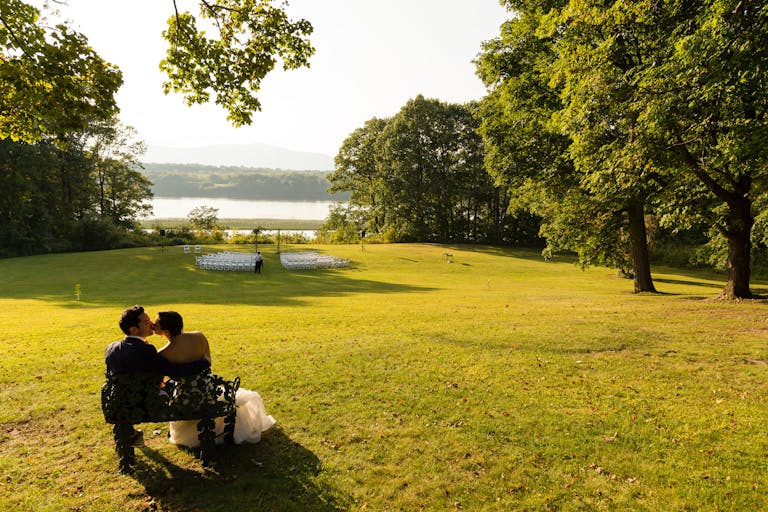 Bride and Groom sitting on a bench outside at their hudson valley wedding venue on the lake | PartySlate