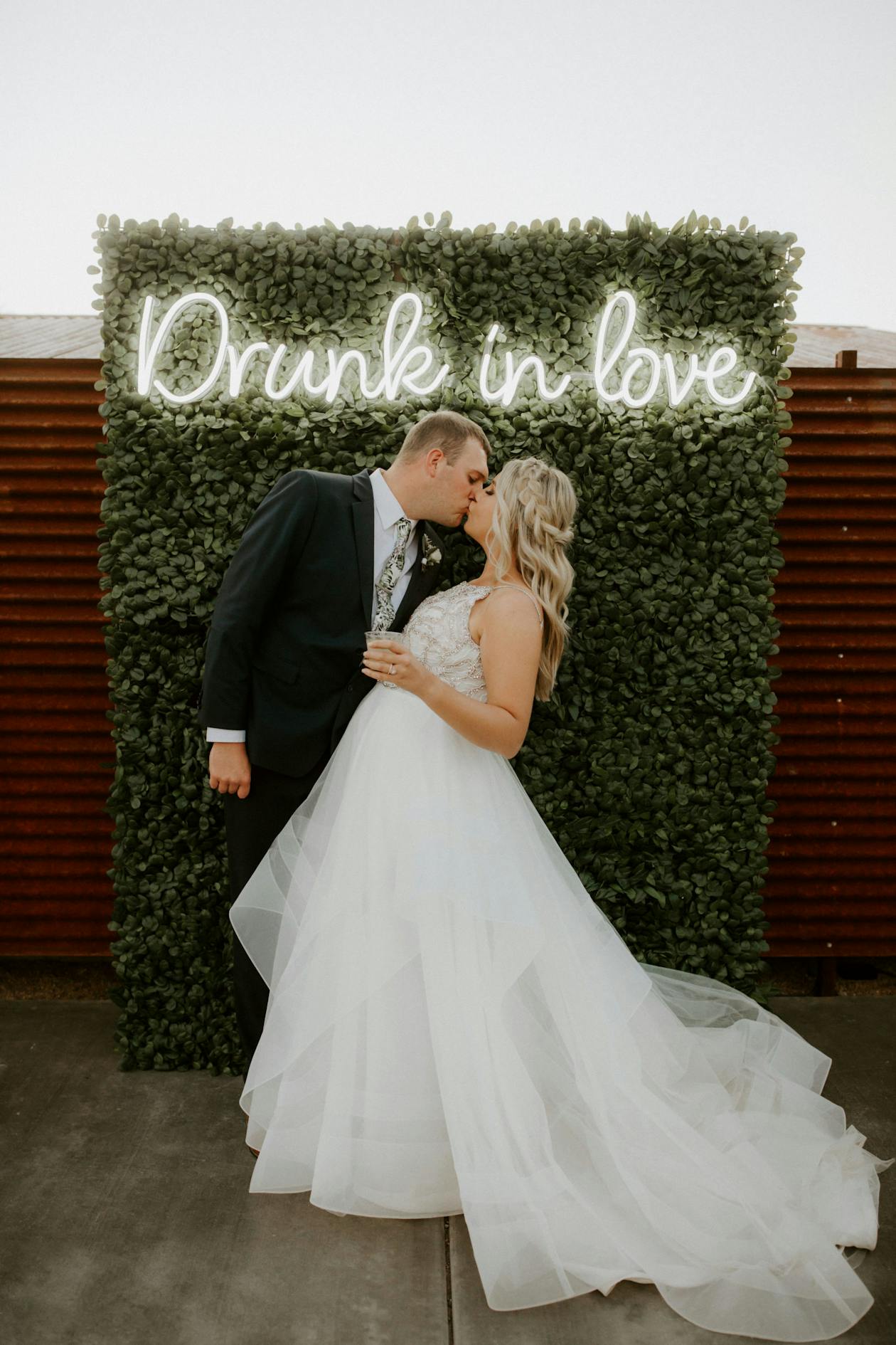 Drunk In Love Neon Sign on Greenery Wall with Couple Posing In front | PartySlate