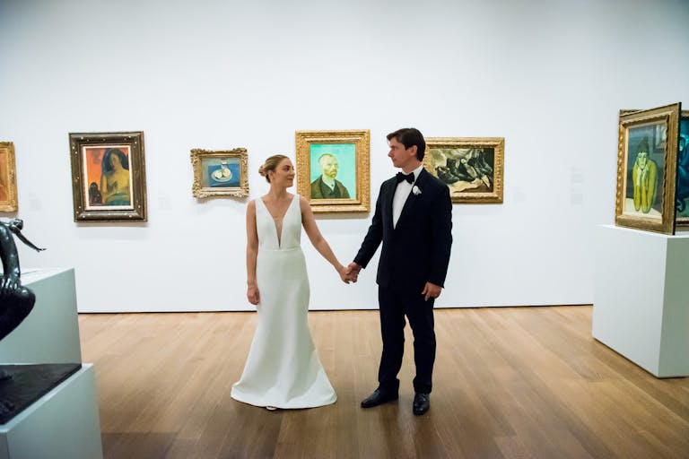wedding Couple holding hands in art gallery wedding venue with art on white walls behind them | PartySlate