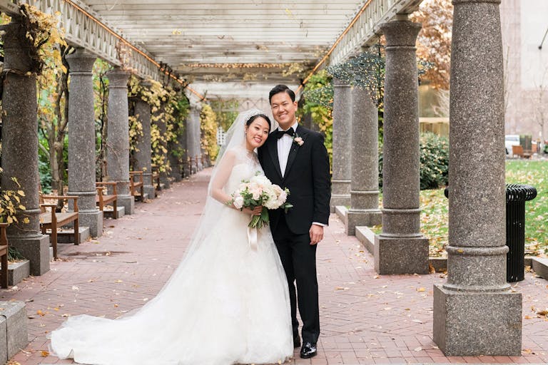 Couple on their wedding day posing and smiling in front of their boston wedding venue with columns lining the pathway | PartySlate