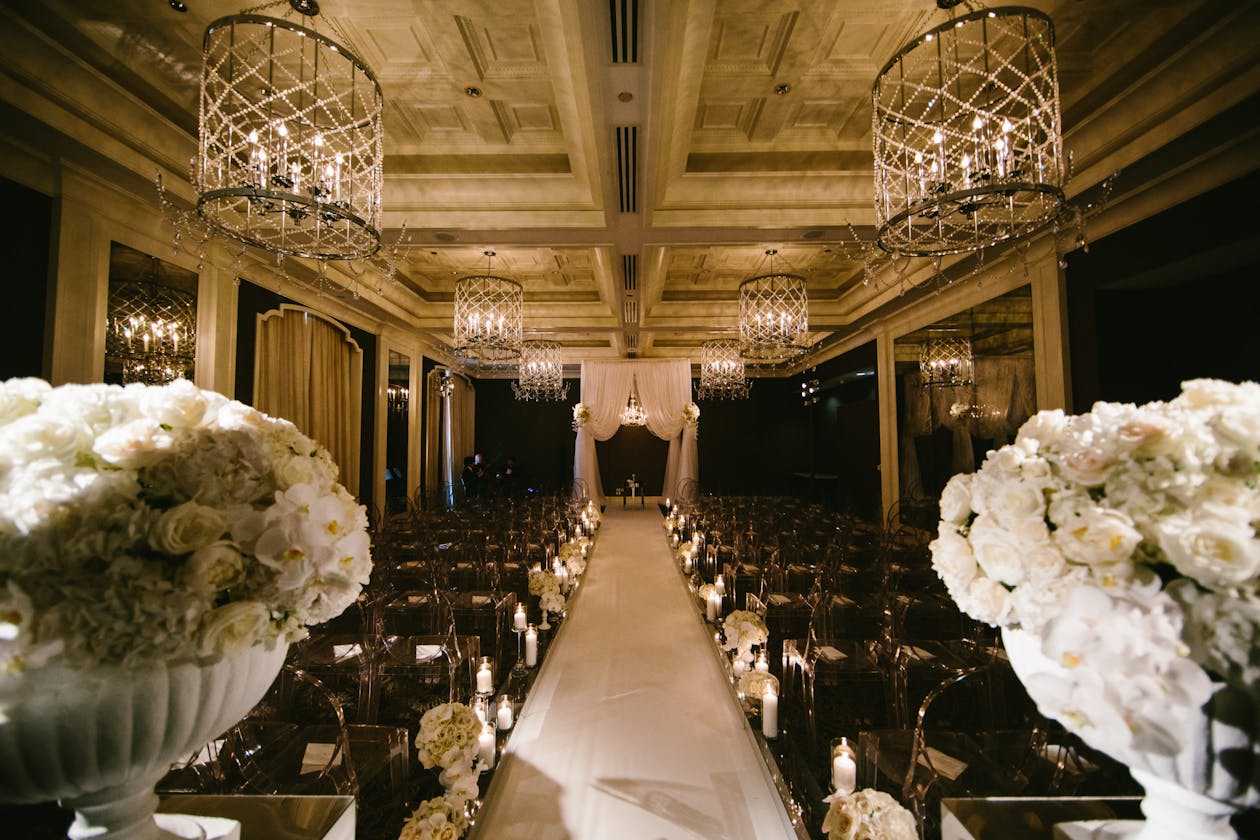 Indoor All White wedding aisle with flowers on either side and floral arch at alter and chandeliers hanging from ceiling | PartySlate