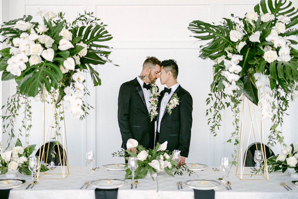 Two grooms touch foreheads at modern sweetheart table with tropical wedding centerpieces | PartySlate