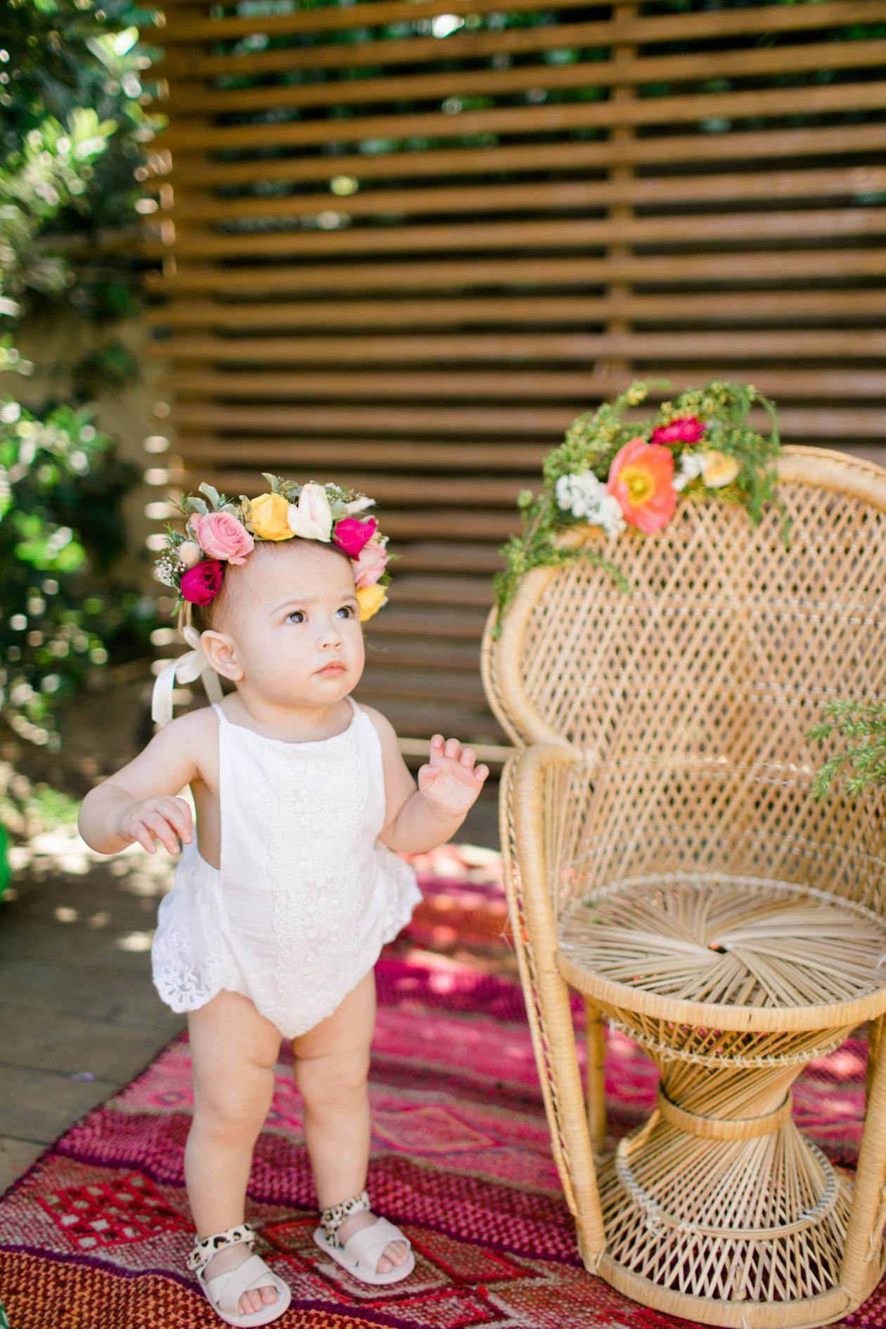 baby with flower crown standing next to whicker chair at her birthday party | PartySlate