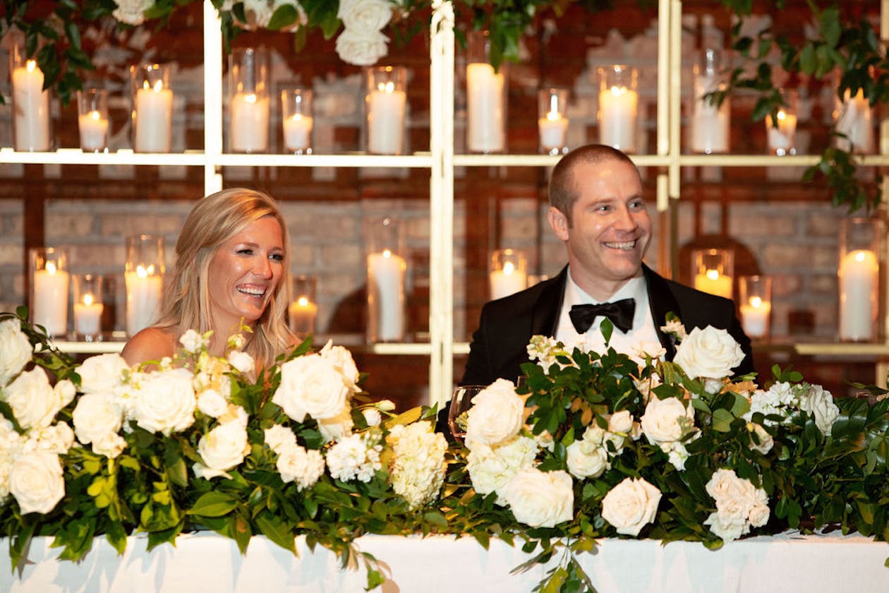 Sweetheart table with the bride and groom sitting and floral centerpiece with greenery | PartySlate