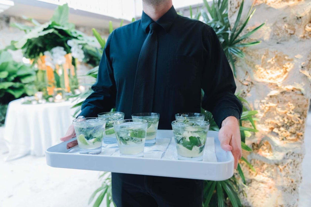 Man in black dress shirt and tie holds white tray filled with cocktails with mint leaves | PartySlate