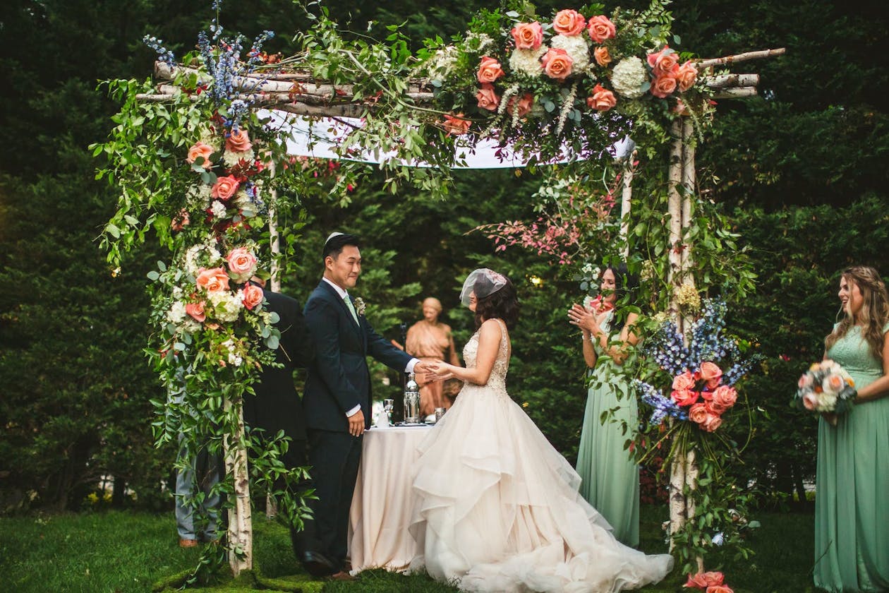 Couple exchange vows under a canopy covered in peach and purple flowers and greenery | PartySlate