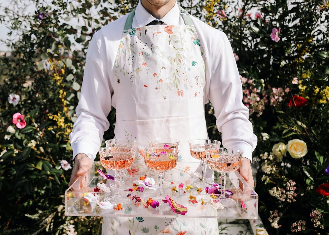 Man in floral apron hold tray with pink cocktails and scattered pink petals | PartySlate