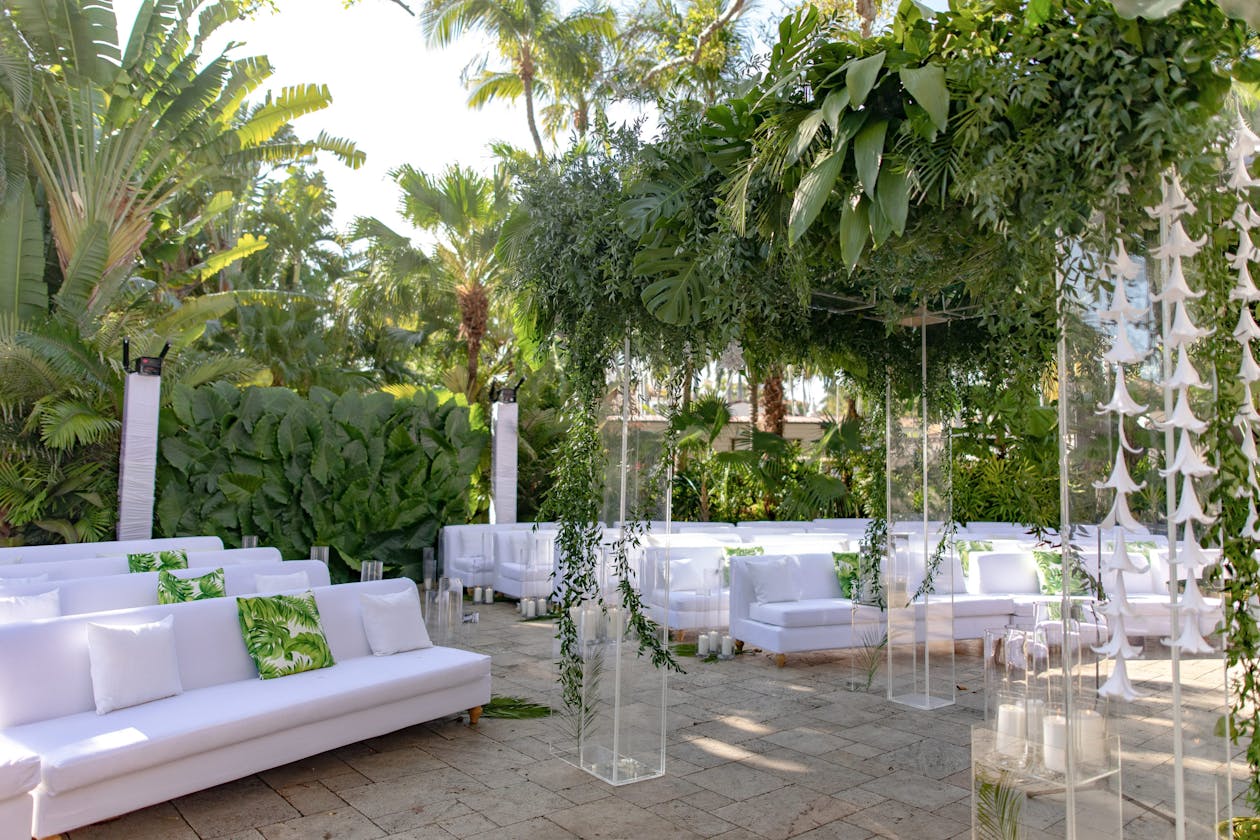 Tropical ceremony with modern white sofas and lush monstera leaves and palm frond foliage | PartySlate