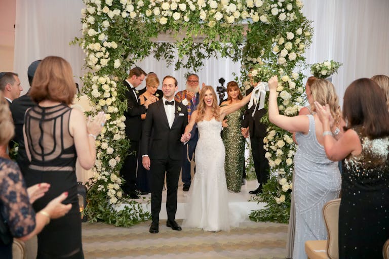 Couple getting married in Boston standing in front of a floral wedding arch inside their venue and holding hands while guests stand and clap | PartySlate