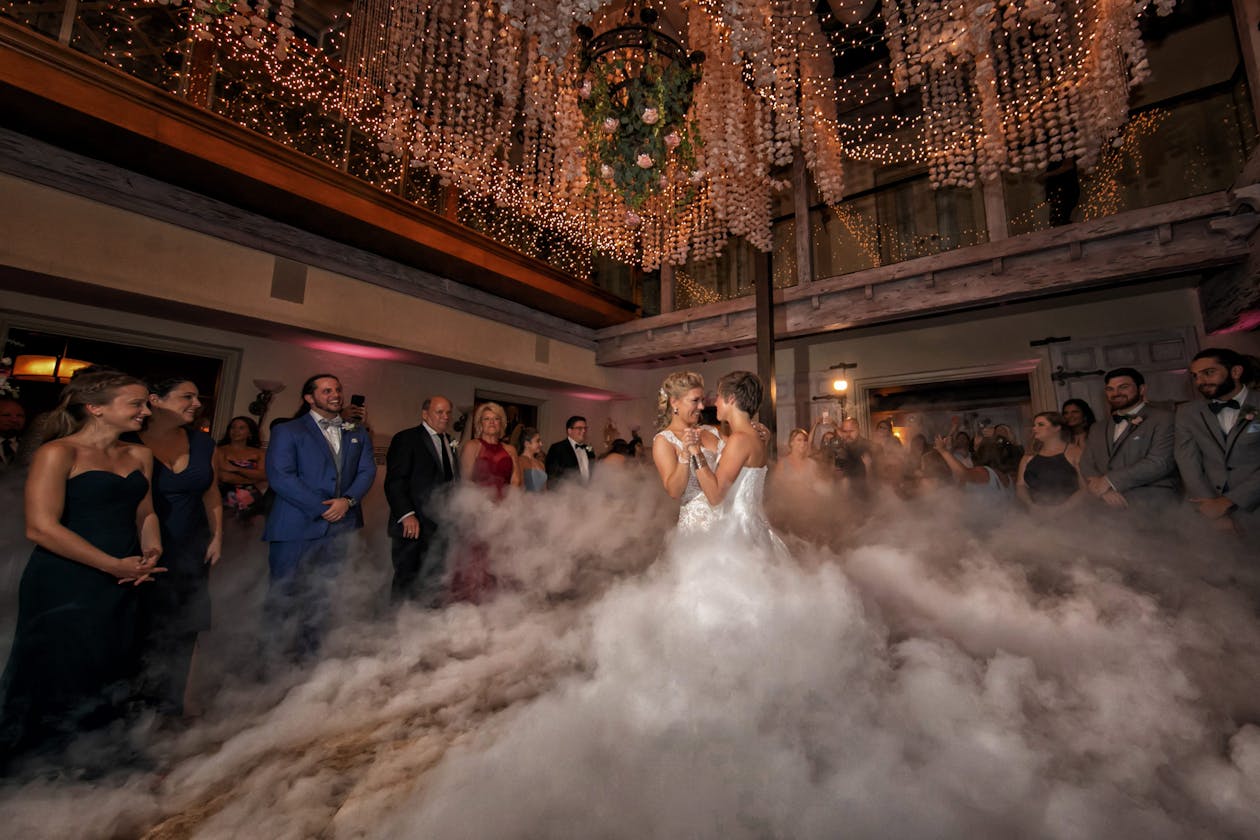 Two brides dance through swirling fog under suspended lights | PartySlate