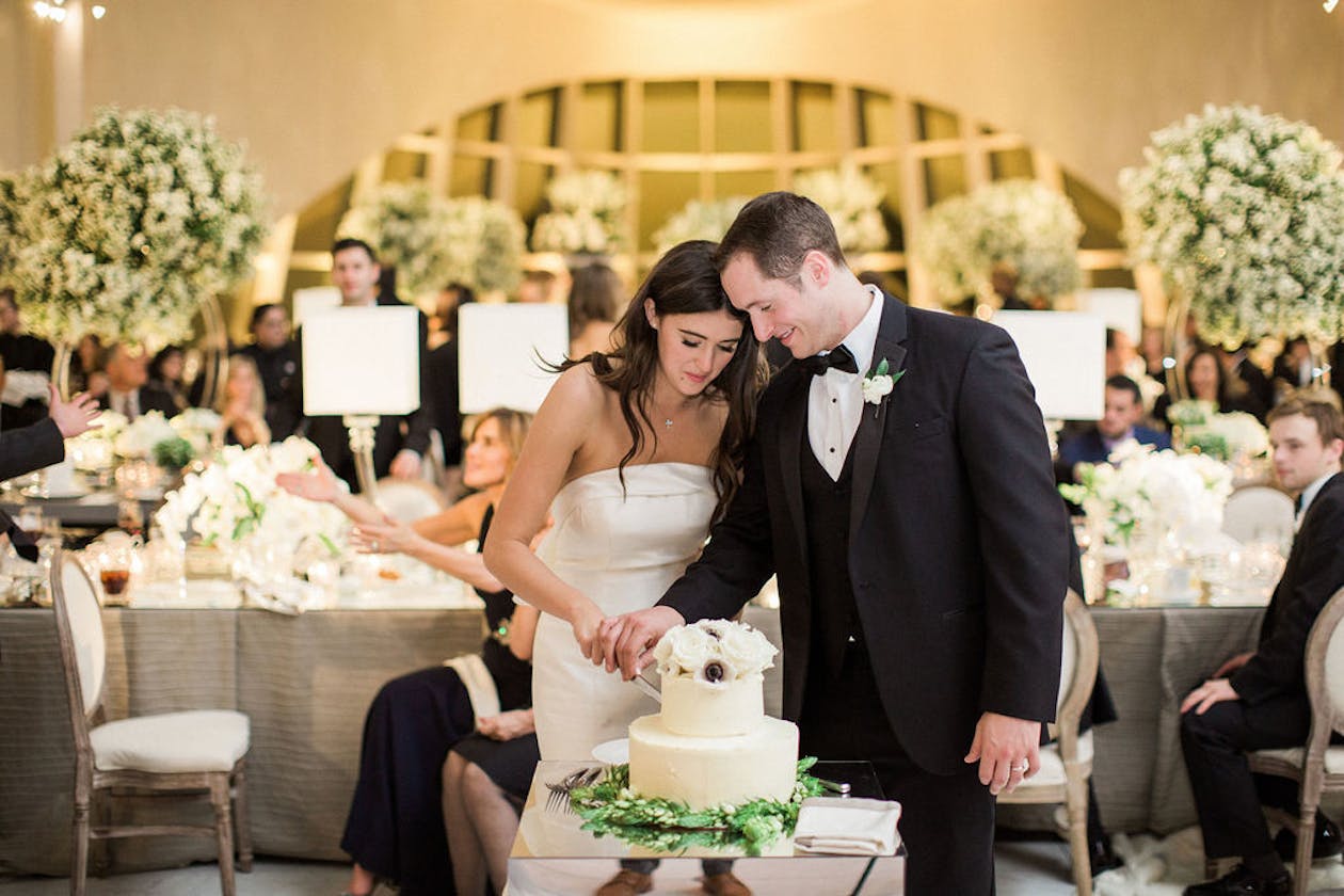 Couple cutting into their white wedding cake with a little bit of greenery on it | PartySlate