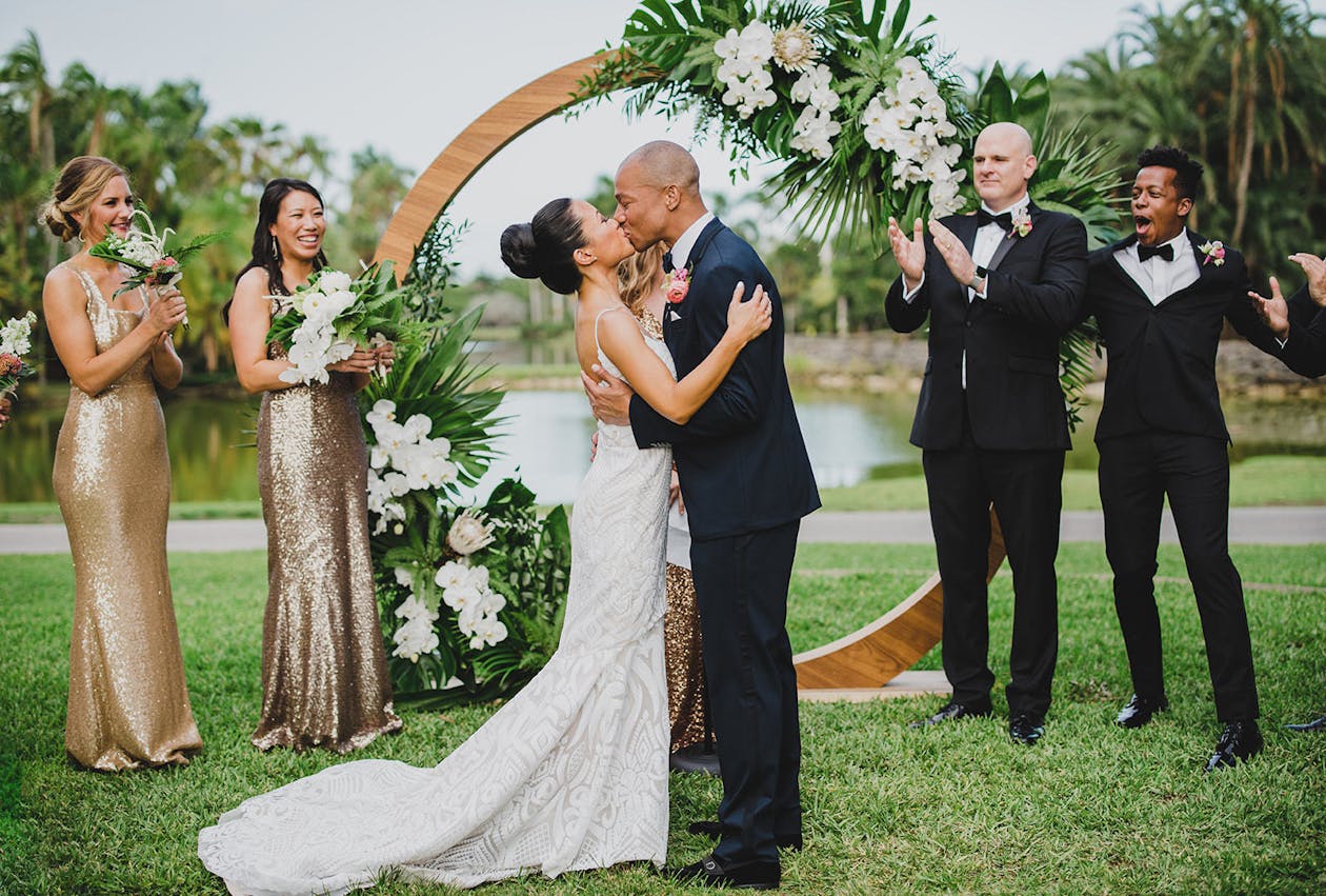 Bride and groom kiss in front of circular gold wedding arch decorated with monstera leaves and white orchids | PartySlate