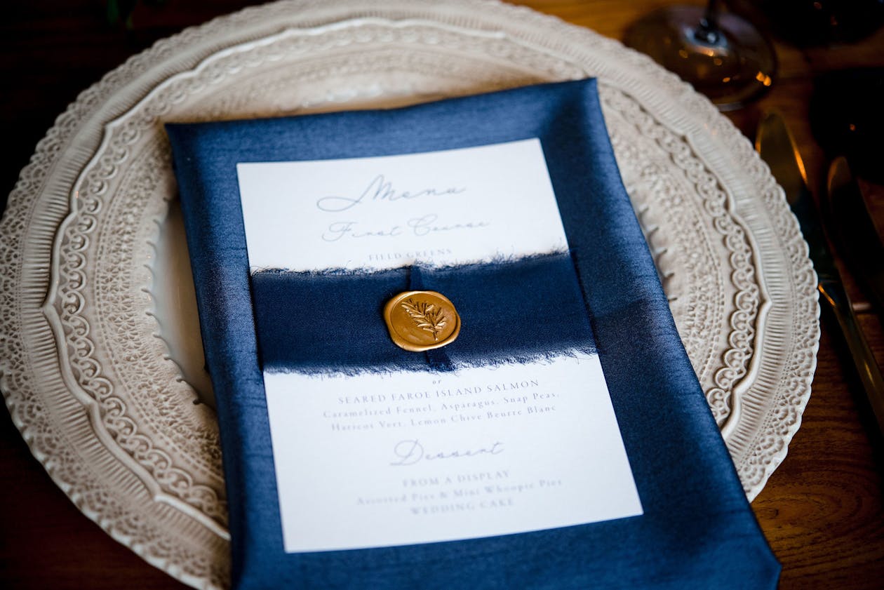 Blue and white wedding invitation with gold seal laid out on navy linen napkin | PartySlate