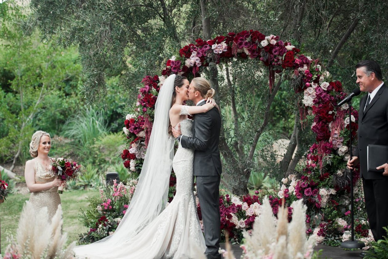 Couple kiss in front of burgundy circular wedding arch | PartySlate