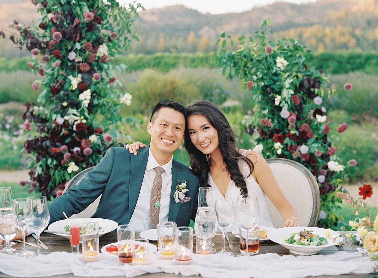 Bride and groom sit at sweetheart table with purple floral and greenery backdrop | PartySlate