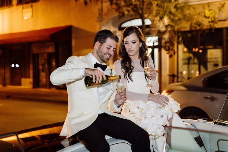 couple popping a bottle of champagne in vintage convertible car on the way to their wedding reception in Miami | PartySlate