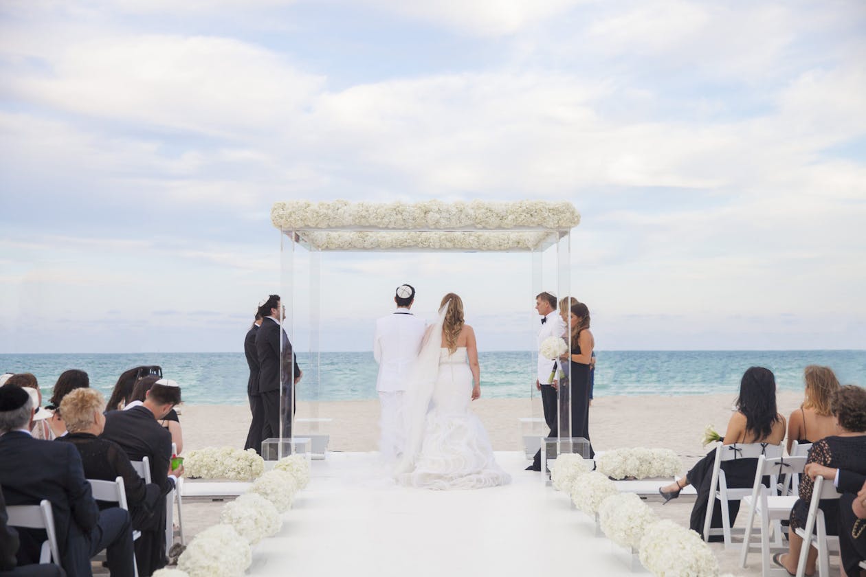 stunning all white wedding on the beach with lucite arch and white florals around it | PartySlate