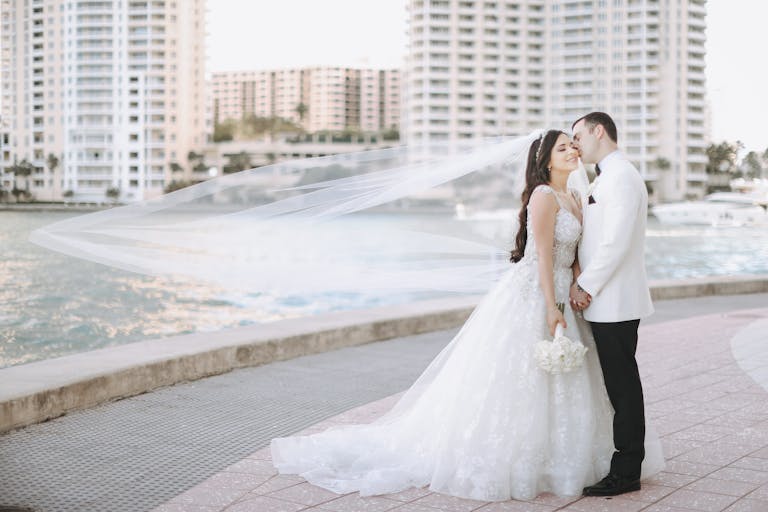 couple standing by body of water with bride's vail blowing in the wind | PartySlate