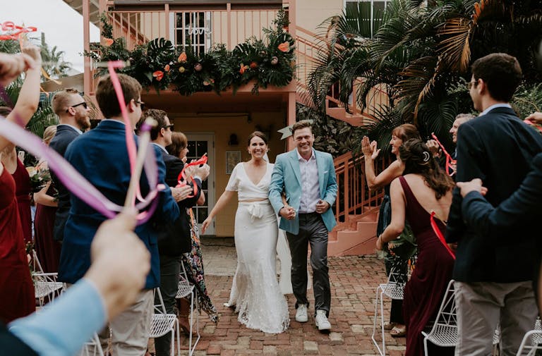 married couple walking down wedding aisle outside with guests waving streamers | PartySlate