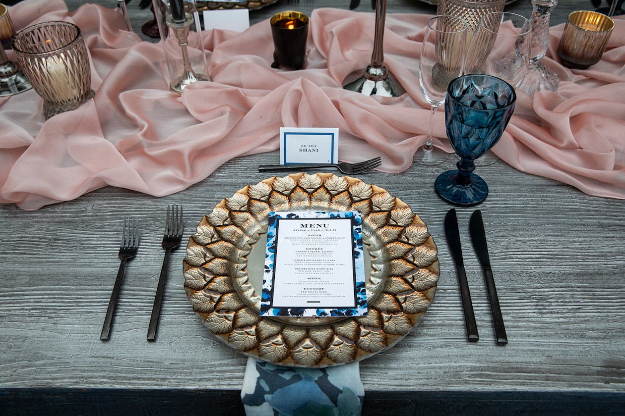 Wedding place setting with gold charger plate, blue goblet, and pink drapery table runner | PartySlate