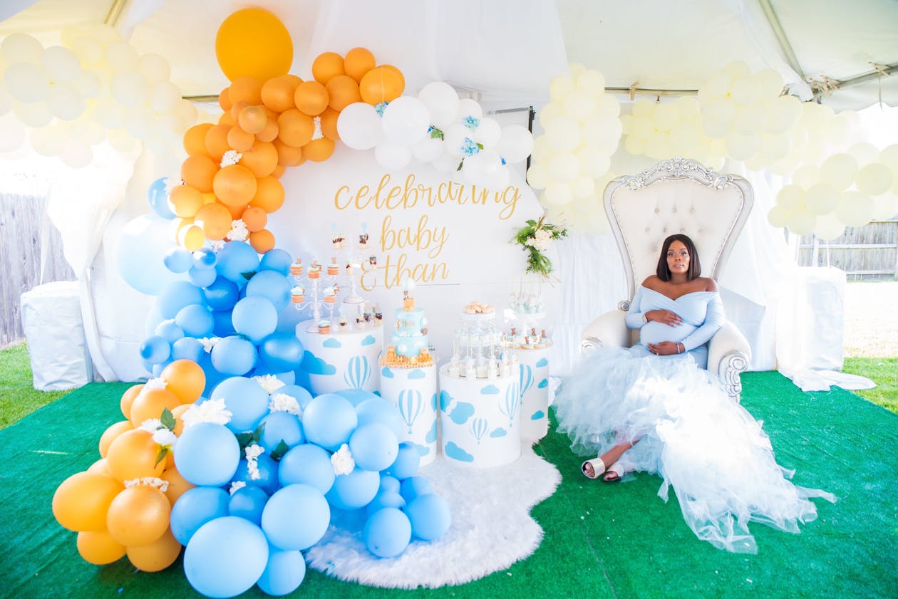 Baby Shower with expecting mother sitting in chair surrounded by blue and yellow balloons and food on stands | PartySlate