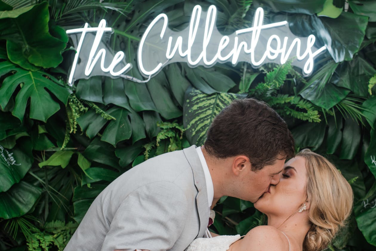 bride and groom kissing in front of greenery wall with neon sign at wedding | PartySlate