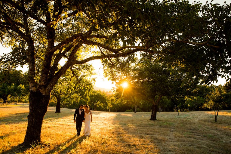 outdoor wedding at triple s ranch in california with couple walking through grass and trees with sun setting behind them | PartySlate
